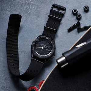 Flat lay of Seiko SRPD79 watch with black ion-coated case, black nylon strap, rotating bezel and day/date on gray background