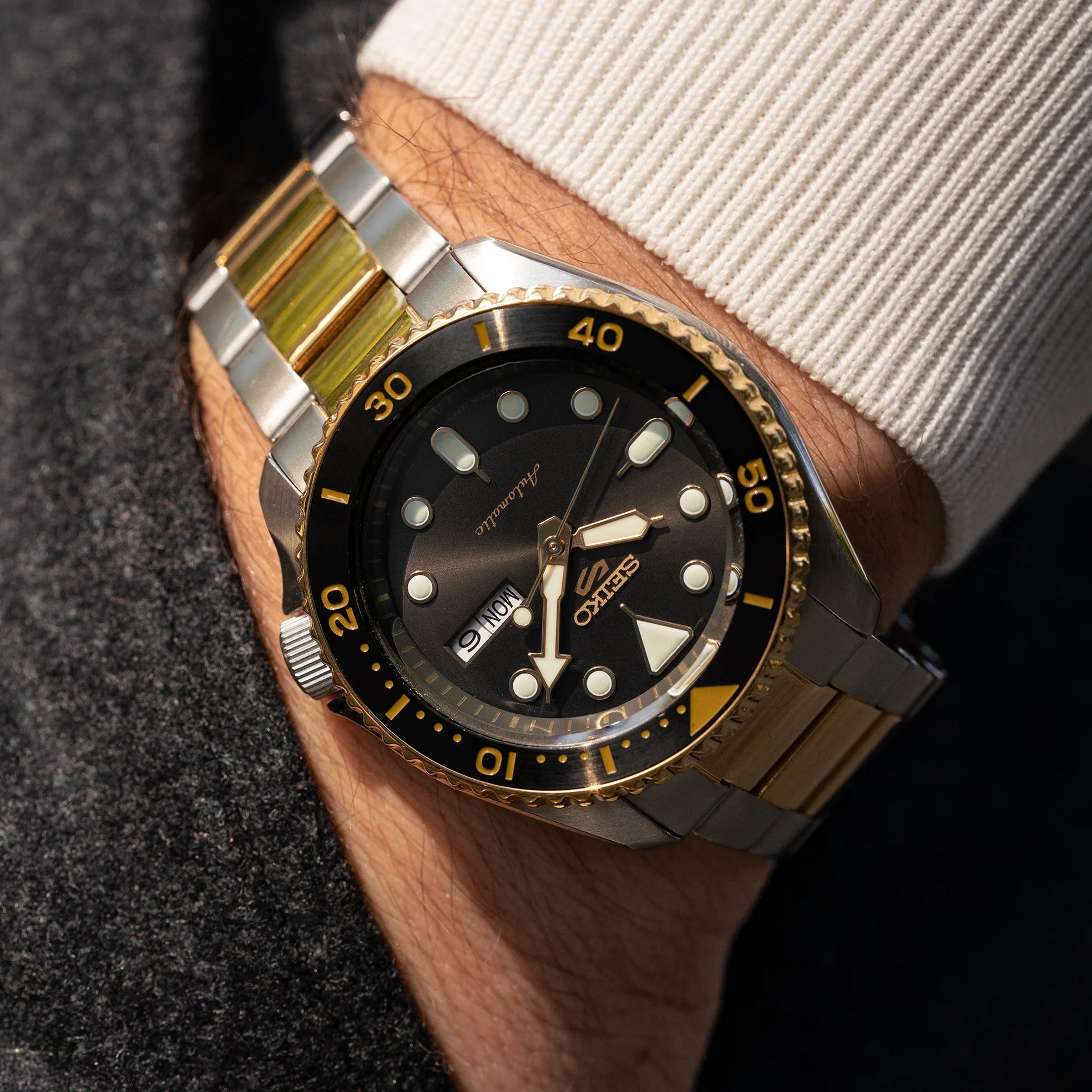 A close-up of a Seiko watch with a two-tone stainless steel and gold-tone link bracelet, worn on a wrist. The watch features a black dial with luminous hour markers, a black rotating bezel with gold accents, and a date window.