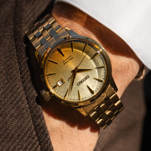 A gold-tone Seiko wristwatch with a metal link bracelet, worn on a person's wrist. The watch has a champagne-gold dial with gold-tone hour markers and hands, a date window at 3 o'clock, and a white dress shirt cuff and brown jacket visible in the background.