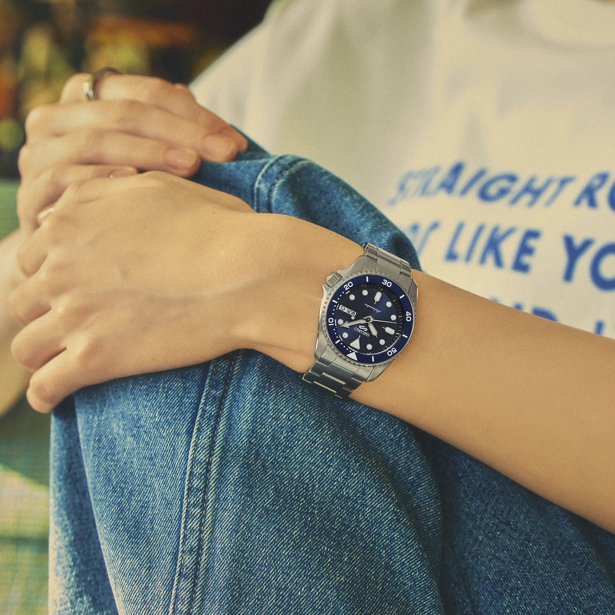 A silver stainless steel wristwatch with a dark blue dial and white markers, worn on a person's wrist. The person is wearing a white graphic t-shirt and blue denim jeans, with their hands crossed over their lap.