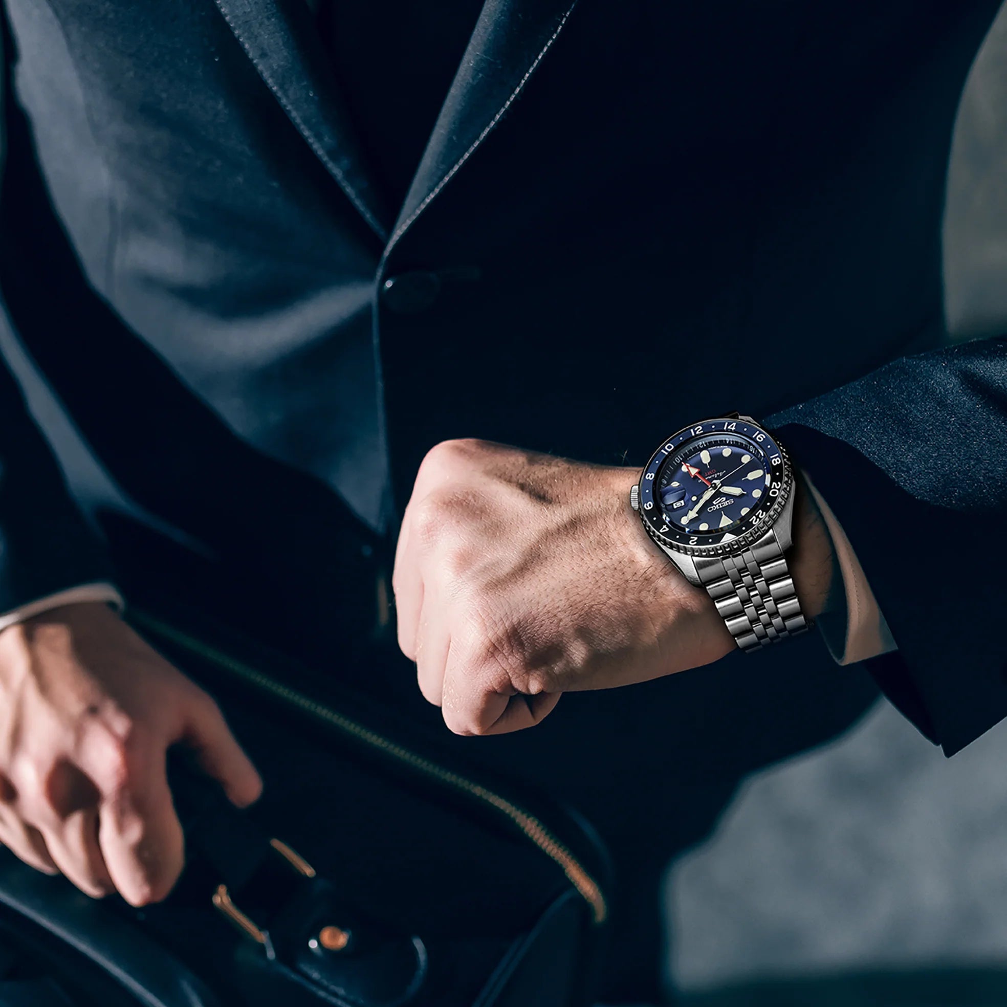 Close-up of Seiko SSK003 watch on wrist showing blue dial, blue-black GMT bezel and stainless bracelet against dark suit