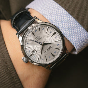 A close-up of a wrist wearing a Seiko analog watch with a silver sunburst dial, black hands and markers, and a dark blue leather strap, set against a background of a person's arm in a brown suit jacket and a light blue patterned dress shirt.