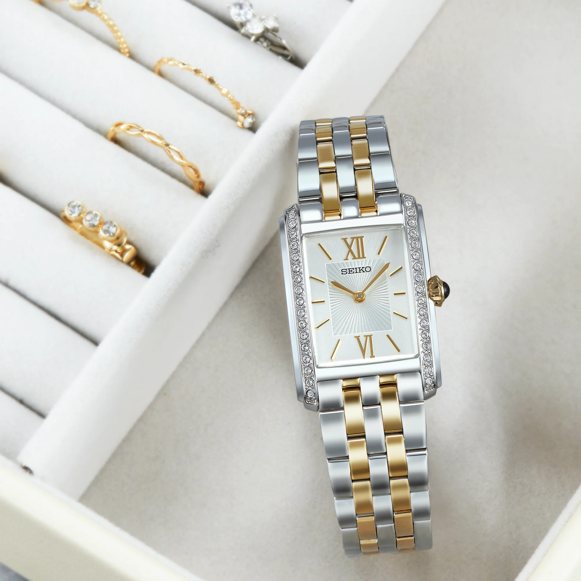 A Seiko women's two-tone watch with a silver and gold metal link bracelet, a white dial with gold Roman numerals, and a bezel accented with small clear crystals, shown next to rings sitting in a beige felt jewelry box