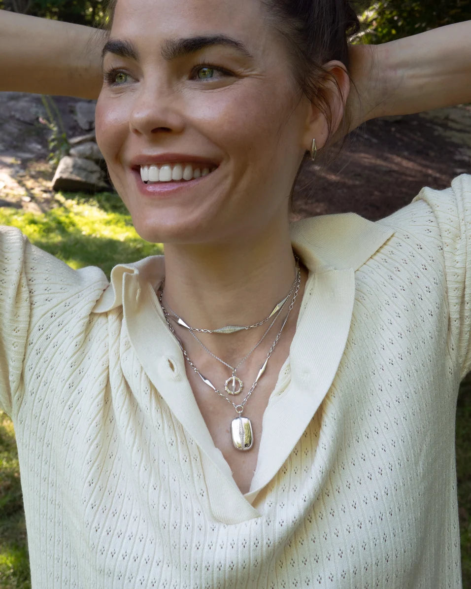 Woman wearing multiple necklaces has her hands behind her head standing outside