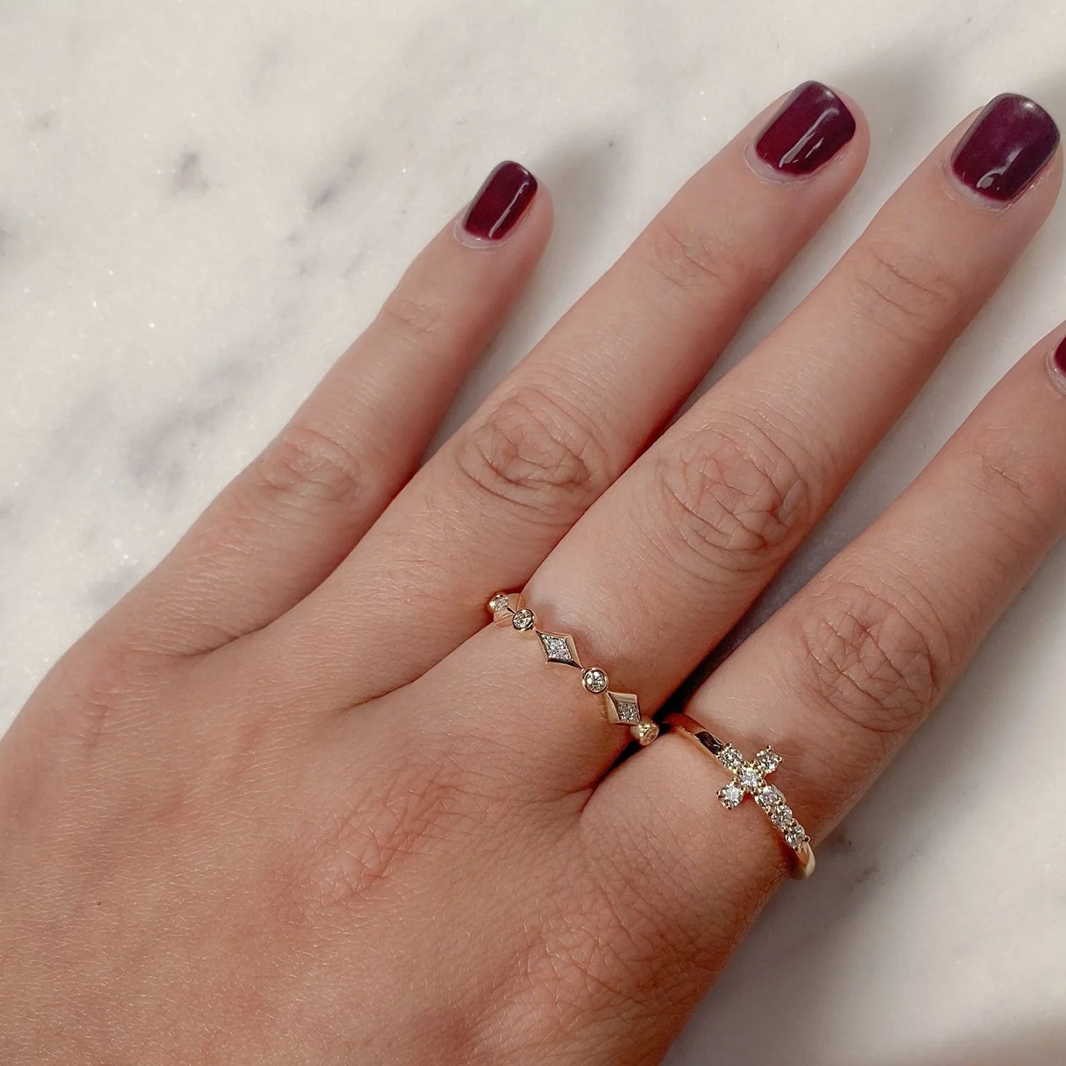 Two thin gold stacking rings on a hand: a diamond cross cluster band and a geometric diamond band.