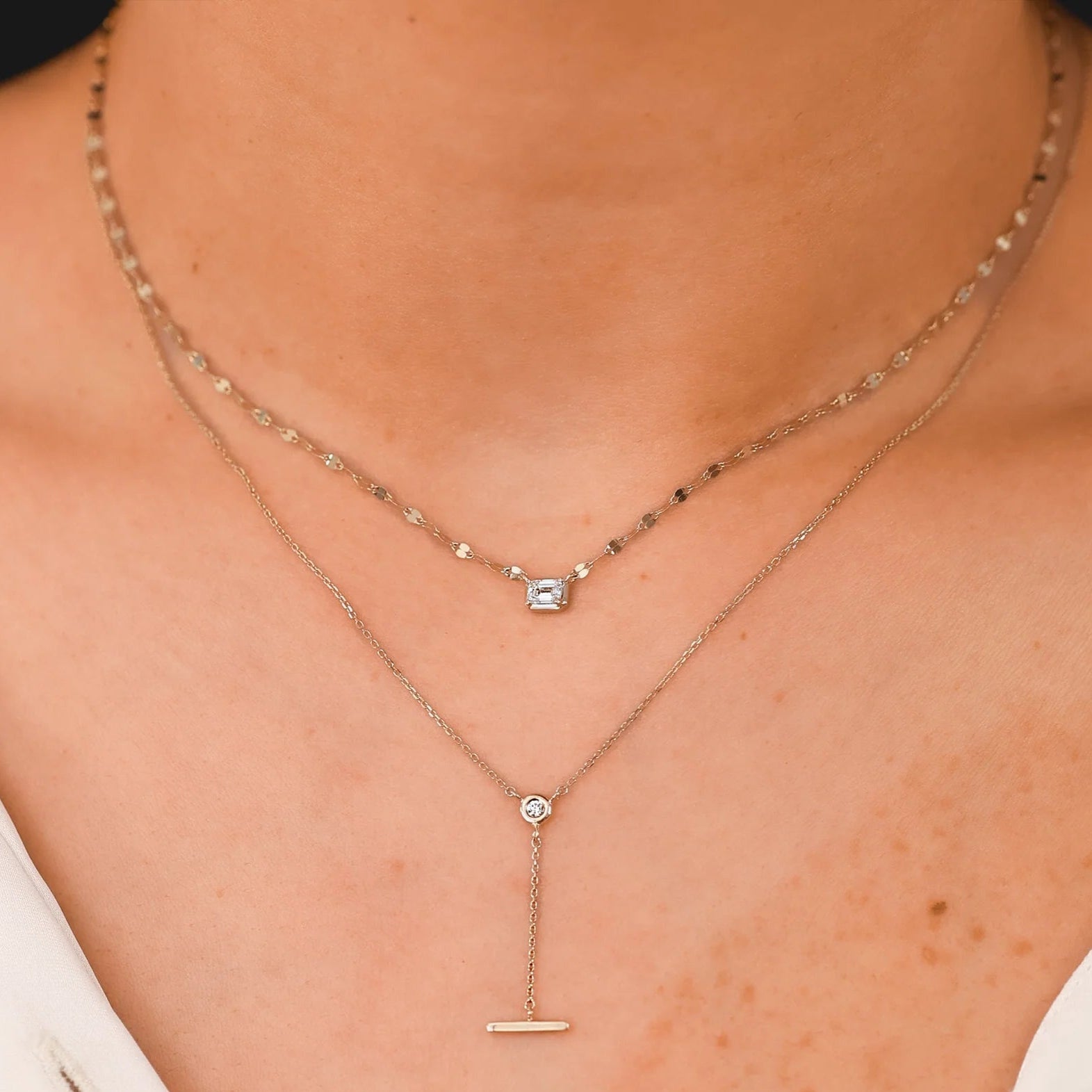 Layered necklaces: emerald-cut diamond pendant on a short chain above a lariat with round diamond and horizontal bar.