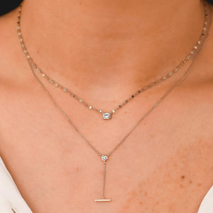 Layered necklaces: emerald-cut diamond pendant on a short chain above a lariat with round diamond and horizontal bar.