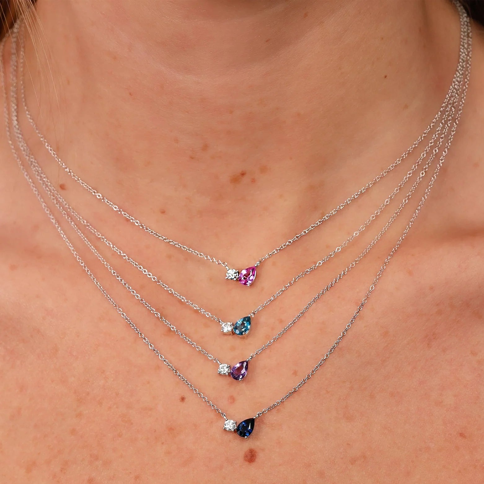 Four-layer silver necklaces on a neck, each with a small round diamond and teardrop gemstone in pink, teal, purple, navy