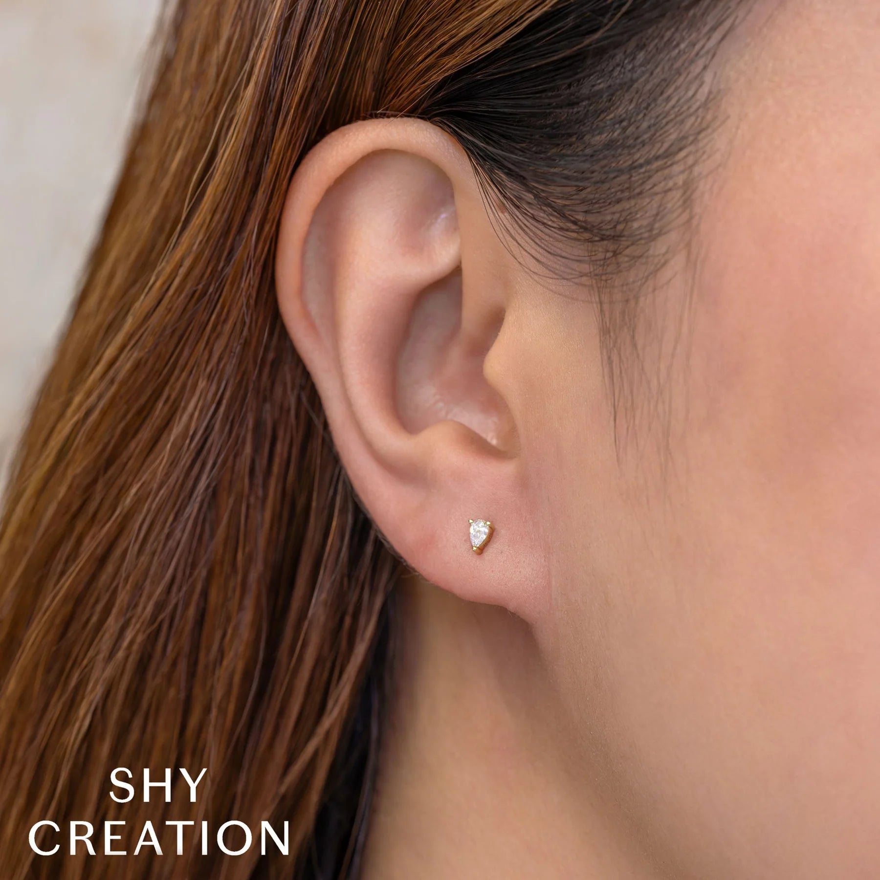 Close-up of earlobe wearing a tiny teardrop diamond stud earring; SHY CREATION logo visible.