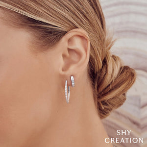 Close-up of a woman's left ear wearing two silver diamond hoop earrings; brand logo SHY CREATION visible.