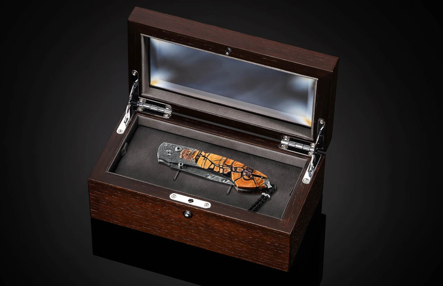 A folding pocket knife with a distinctive orange and black wood grain handle is displayed inside an open dark brown wooden presentation box with a glass lid, set against a dark background.