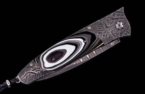 A folding pocket knife with a dark gray Damascus steel pattern handle and a black, white, and gray concentric eye-like inlay on the blade, shown against a solid black background.