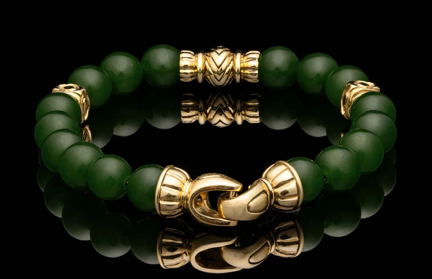 A beaded bracelet made of round dark green jade beads with polished gold-toned metal spacers and a lobster clasp, displayed on a glossy black reflective surface.