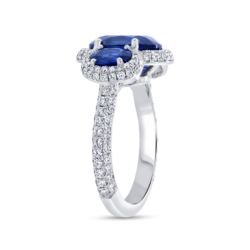 Angled side view of three-stone oval blue sapphire ring showcasing diamond halo and pave diamond band on white background