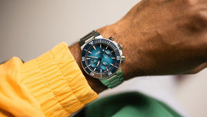 Close-up of a wrist wearing a watch with a blue face and stainless steel strap, against a neutral background.
