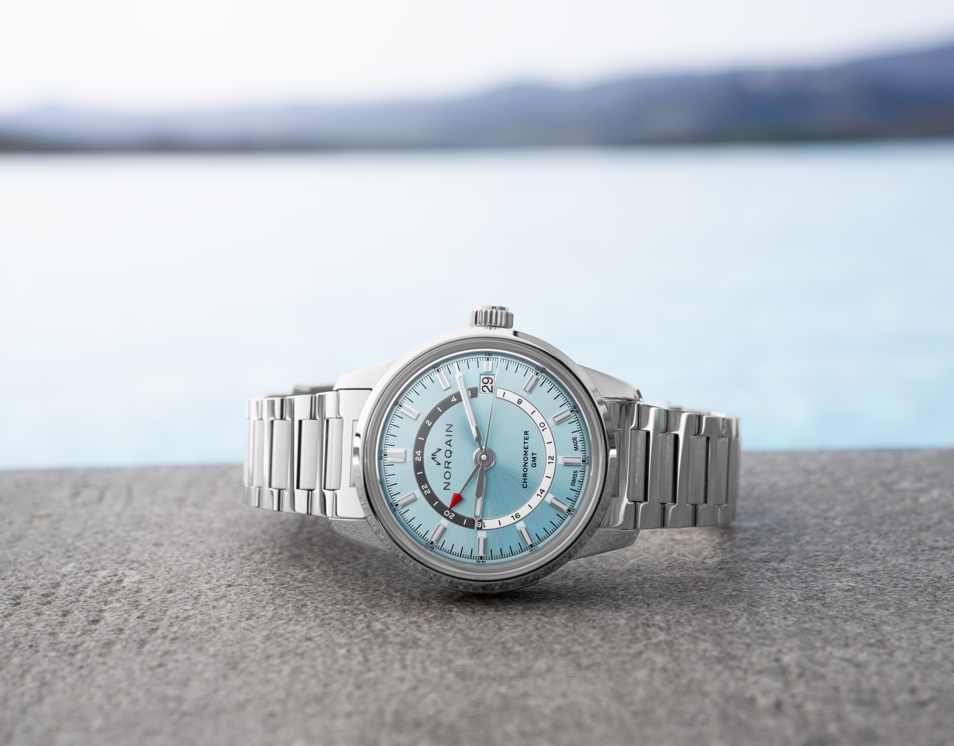 Front view of Freedom 60 GMT 40mm watch with blue dial, red-tip GMT hand, date at 3 o'clock, stainless bracelet and blurred sea