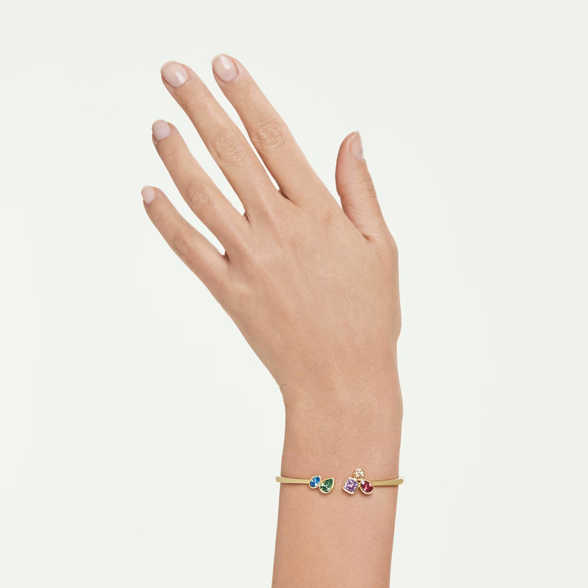 a hand and write wearing a Gold tone bangle with multicolored stones with different cuts on a white background.