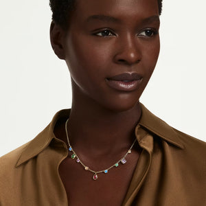 woman wearing gold shirt along with a gold necklace with multicolored stones  on a white background