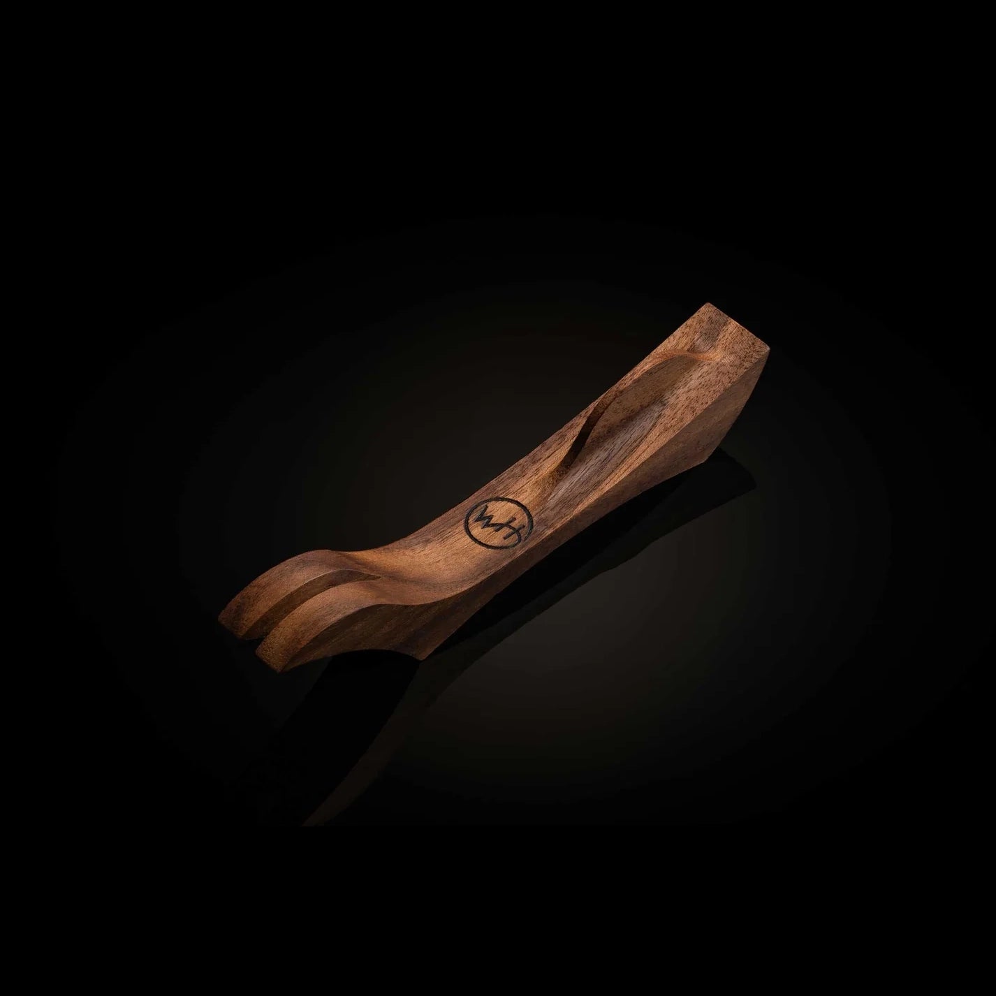 A carved wooden spoon or scoop with a small circular logo, shown on a dark reflective background.