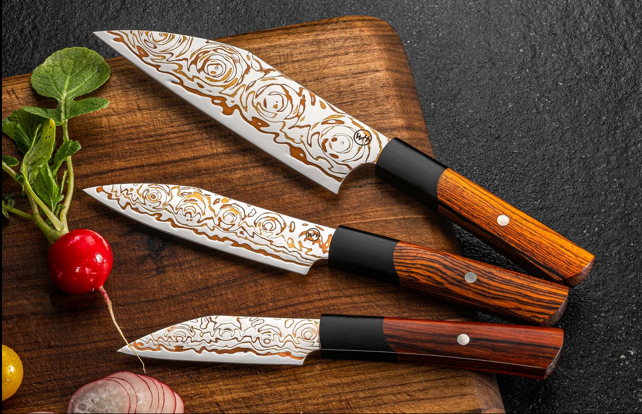Three patterned chef knives with wood handles on a wooden cutting board beside radish and sliced onion