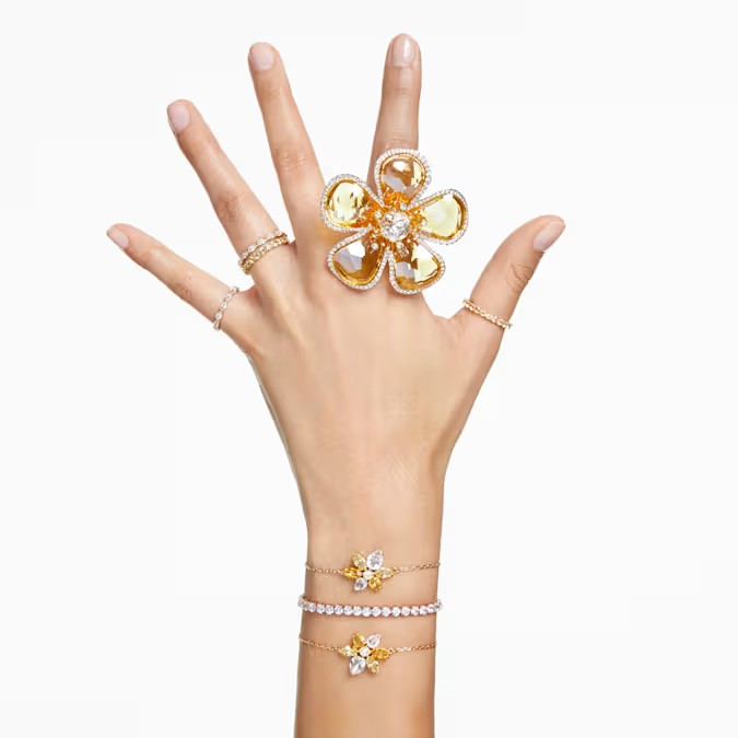 Woman's hand displaying oversized yellow flower cocktail ring, three gold bracelets and several delicate stackable rings on white background