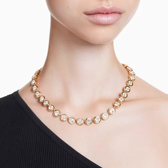 Front view of Imber Tennis Necklace on model, gold-tone plated with bezel-set round crystals, worn with black one-shoulder top