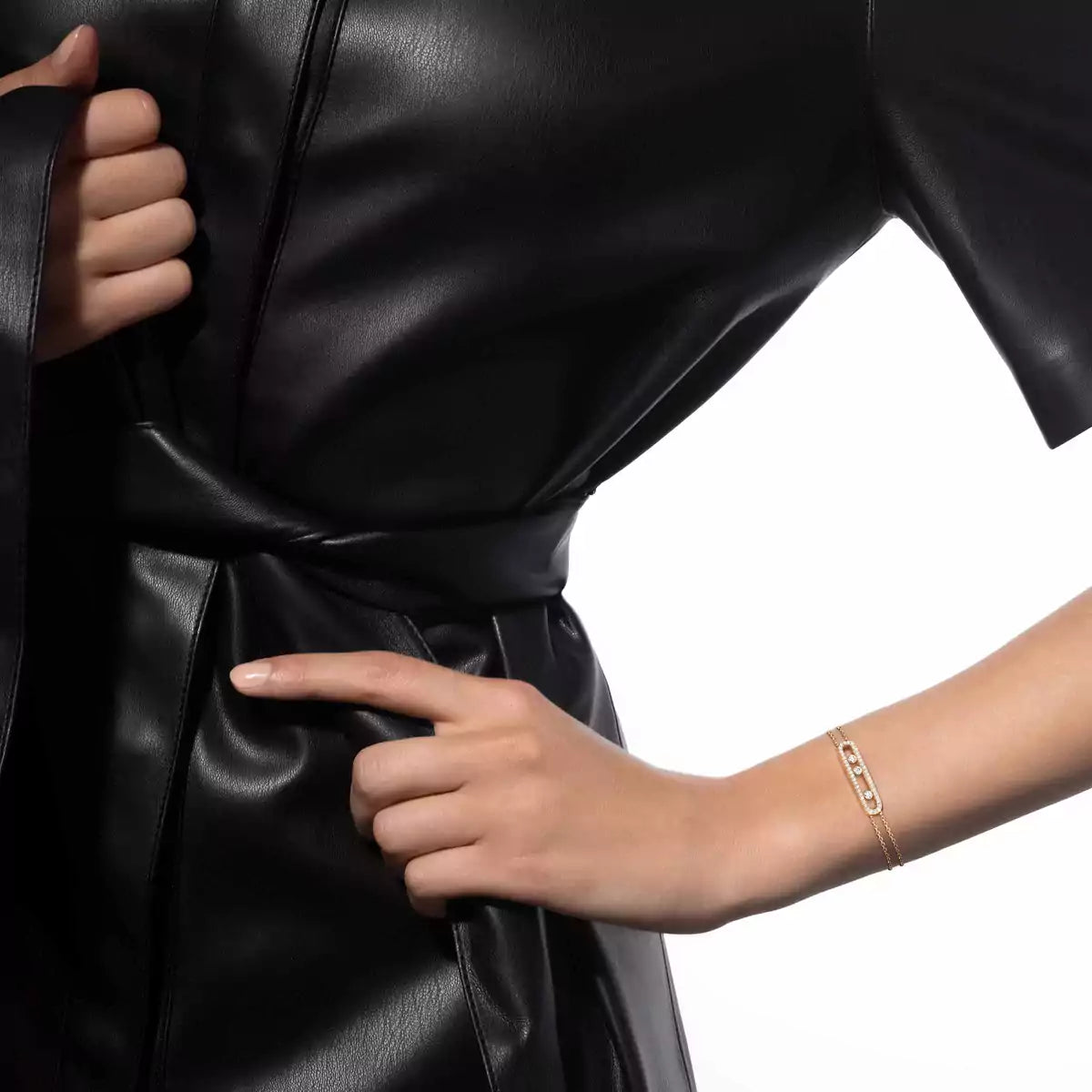 Close-up of a person adjusting the waistband of a black leather outfit on a white background wearing a gold bracelet