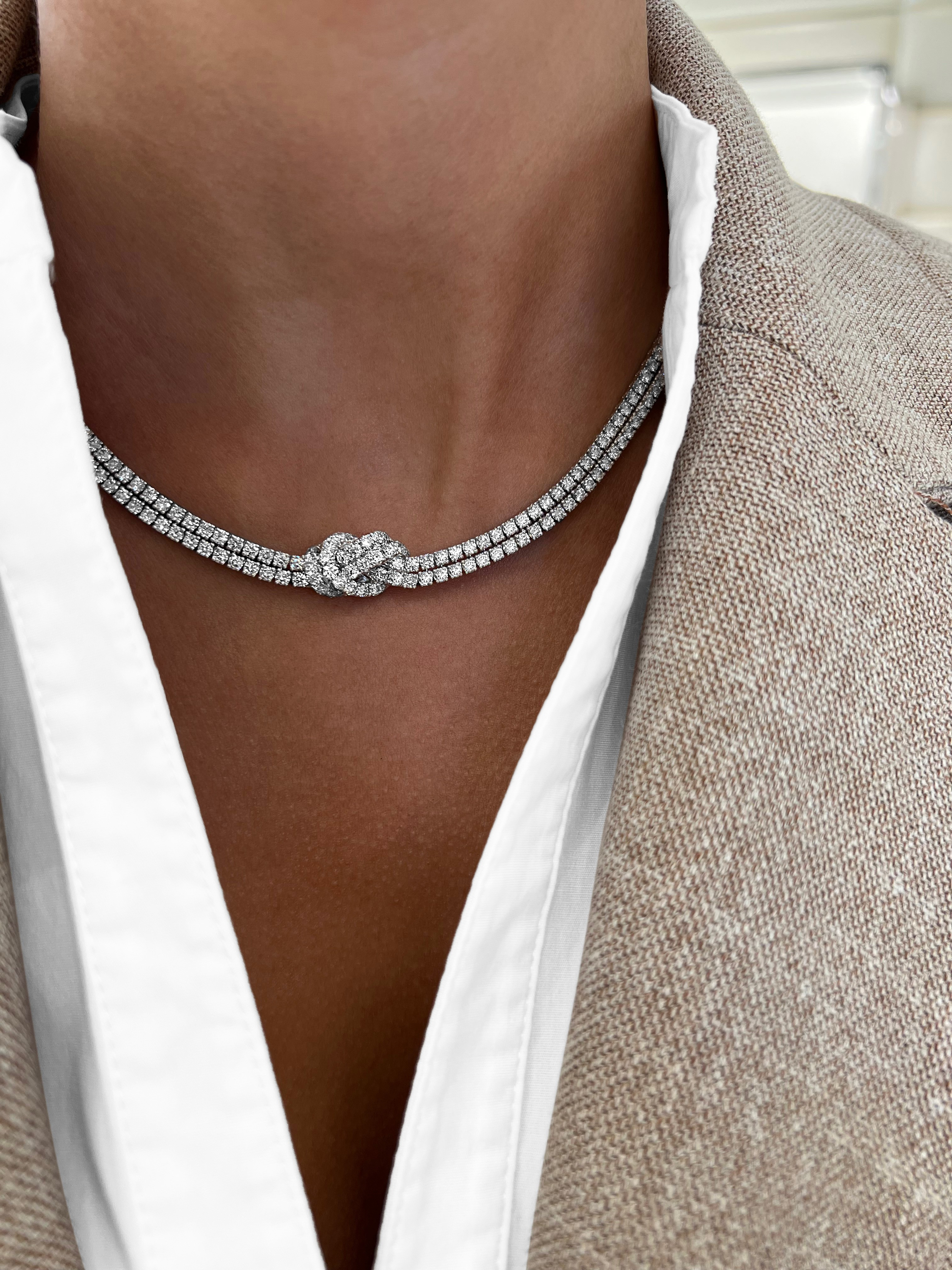 A close-up of a model wearing a silver-toned diamond choker necklace with a knot design at the center, styled with a white collared shirt and a beige blazer.