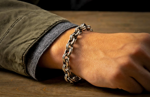 Silver rope-link bracelet on a wrist resting on a wooden table, olive jacket cuff visible