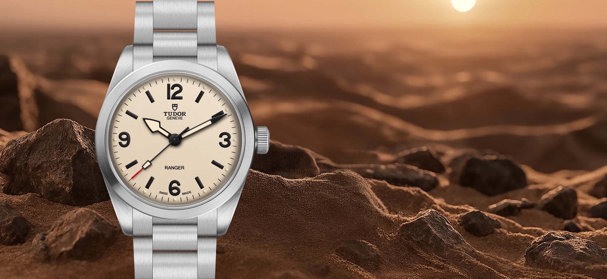 A Tudor Ranger watch with a silver stainless steel bracelet and a cream-colored dial, set against a rocky desert landscape at sunset.