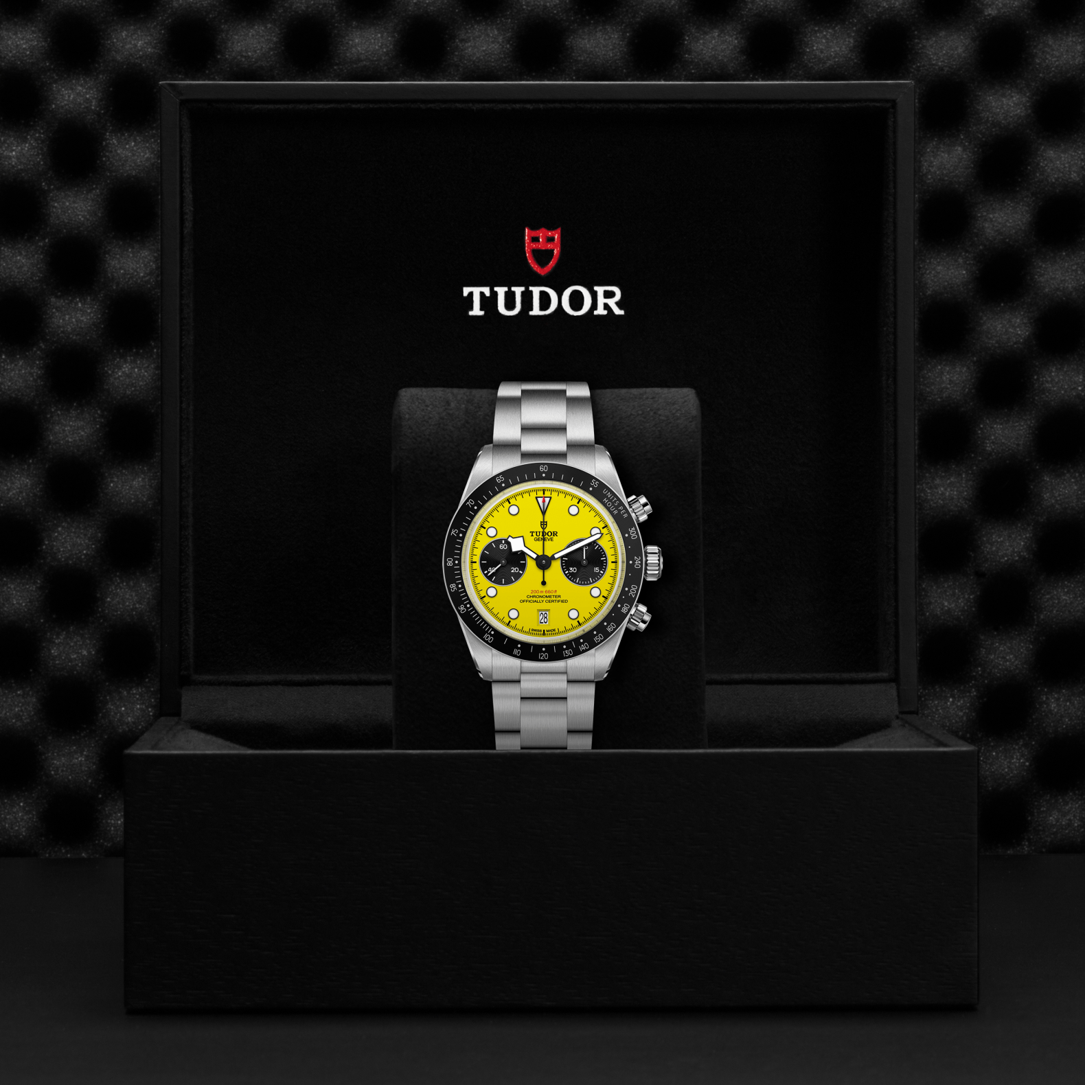 Tudor watch with yellow face and silver bracelet on a black display stand against a textured black background.