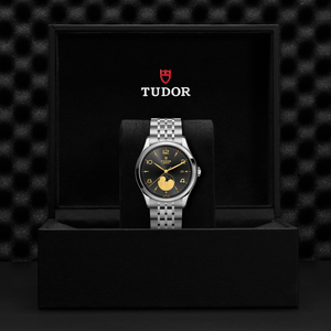 Front view of TUDOR 1926 Luna watch with black sunburst dial, gold numerals and moon phase, on steel bracelet in black Tudor box