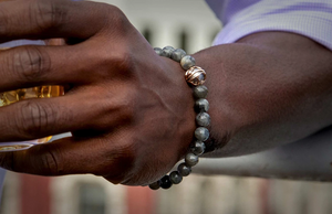 Close-up of wrist wearing gray stone beaded bracelet with a gold decorative bead, hand holding a tumbler of amber liquid.