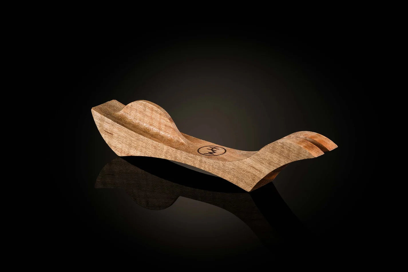 A handcrafted wooden pocket knife rest with a curved, sculptural design, featuring a natural light brown wood grain and a small circular logo in the center, set against a dark reflective background.