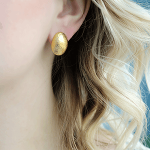 Close-up of left ear wearing 24K gold 18mm stud earring showcasing hammered dome texture with blurred light-blue background