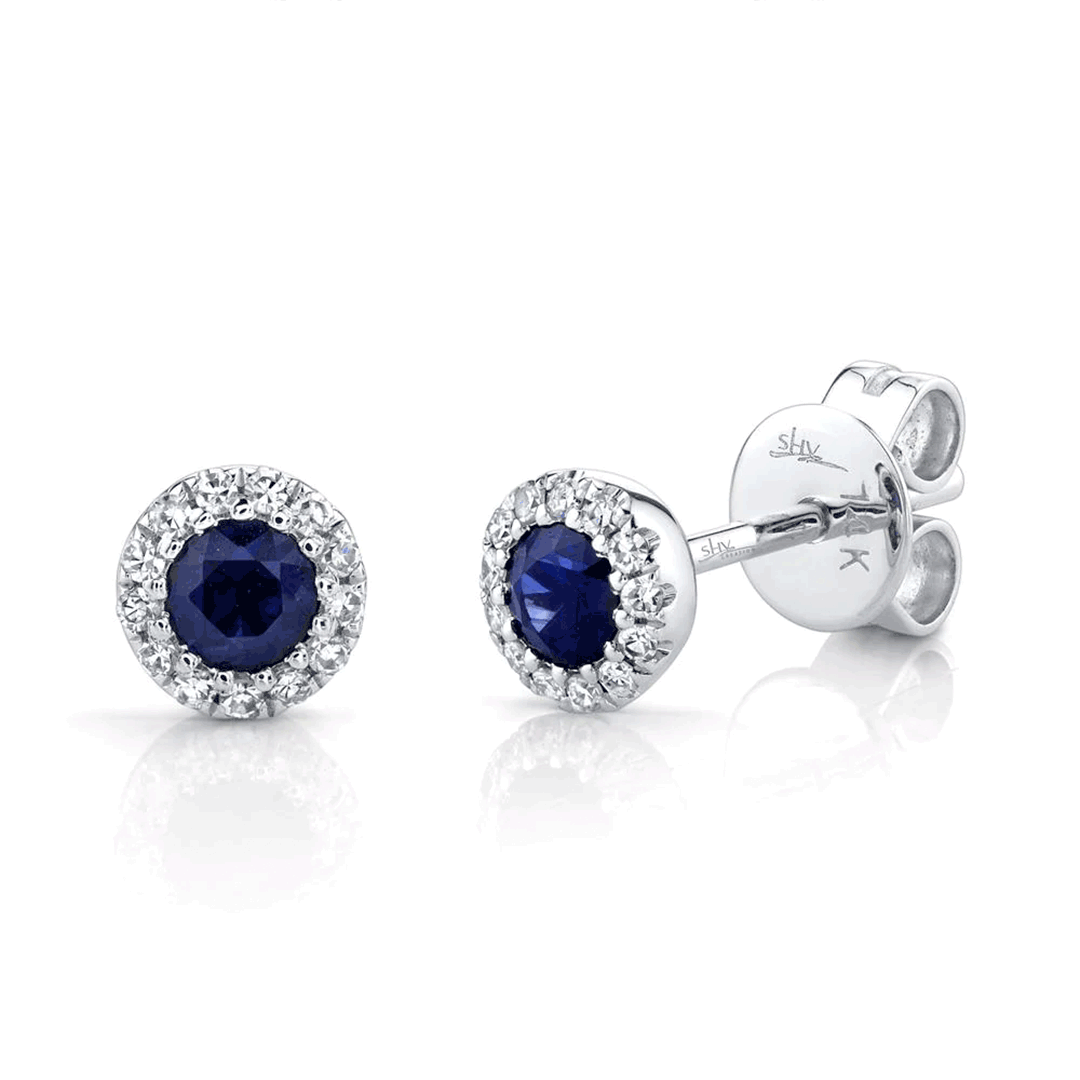White-gold stud earrings with round blue sapphires surrounded by diamond halos; hallmarks 'shv' and 'K' visible on the post.