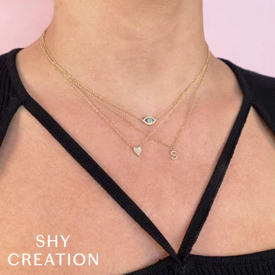 Three layered gold necklaces with an evil eye, small heart, and 'S' initial charms; text 'SHY CREATION' lower-left.
