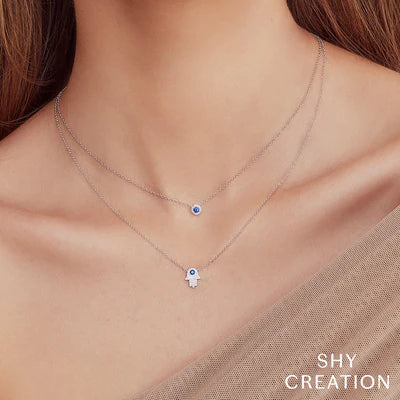 Delicate double-layer silver necklaces with a blue evil-eye pendant and Hamsa hand charm; SHY CREATION logo visible.