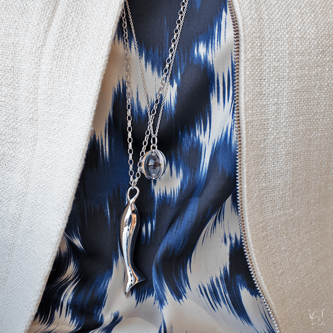 Perseverance fish charm necklace with two silver chains draped over blue patterned fabric and a cream jacket zipper