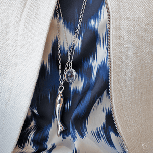 Perseverance fish charm necklace with two silver chains draped over blue patterned fabric and a cream jacket zipper